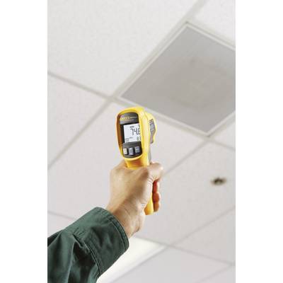 Eine Person hält ein gelbes Infrarot-Thermometer und misst die Deckenoberflächentemperatur in einem Büro. Display zeigt '74,8 F'.