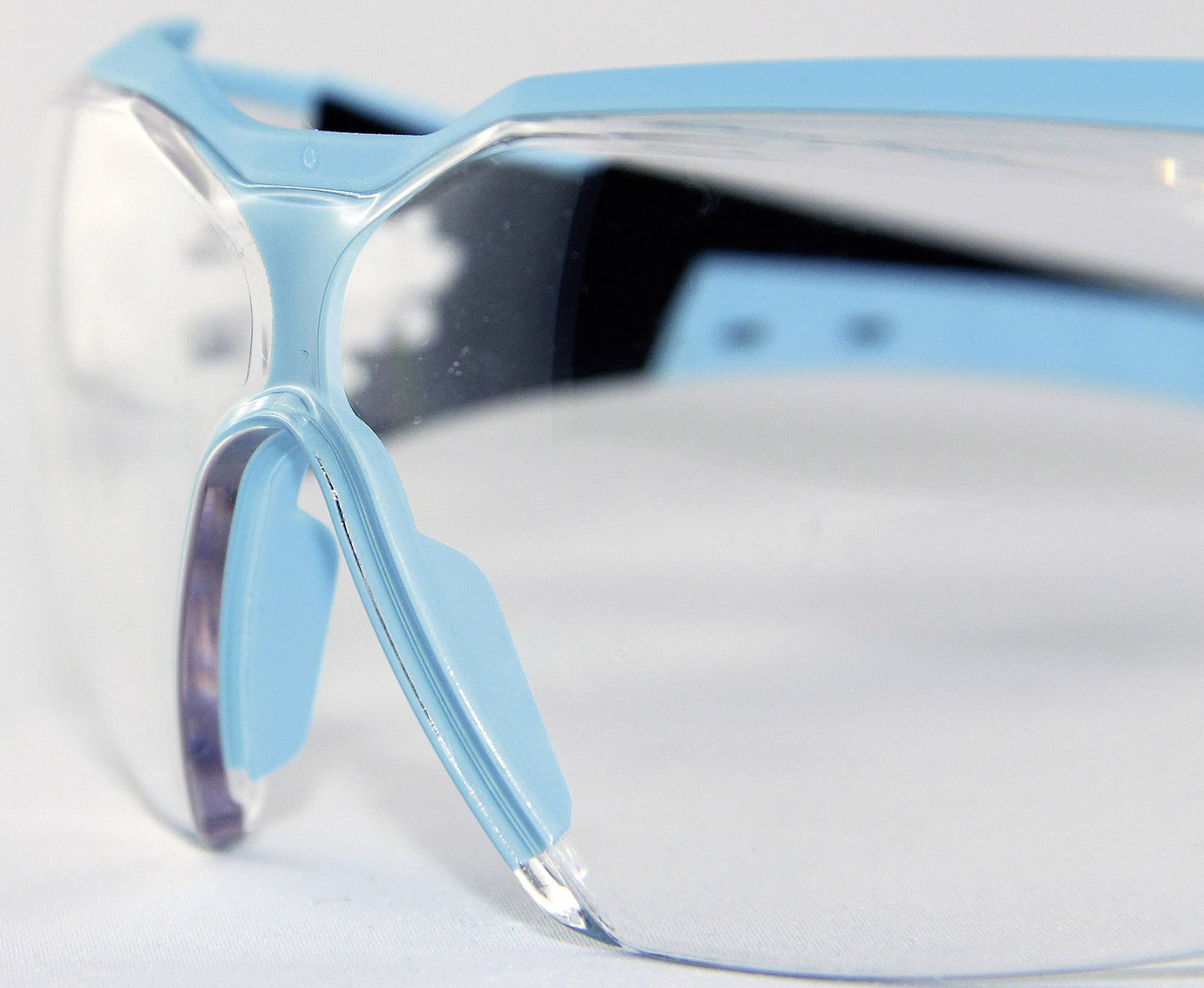 Eine hellblaue Schutzbrille mit transparenten Gläsern und breiten, schwarzen Bügeln liegt auf einer weißen Oberfläche.
