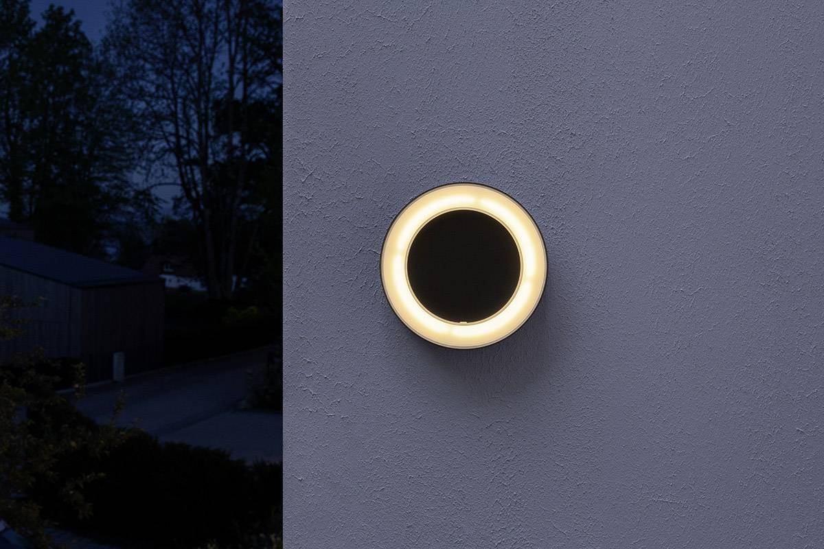 Eine runde, leuchtende Außenlampe an einer grauen Wand, bringt warmes Licht in eine dunkle Umgebung.