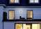 Ein modernes Haus mit einer verglasten oberen Terrasse. Zwei Stühle sind dort platziert. Die Fenster strahlen warmes Licht aus.
