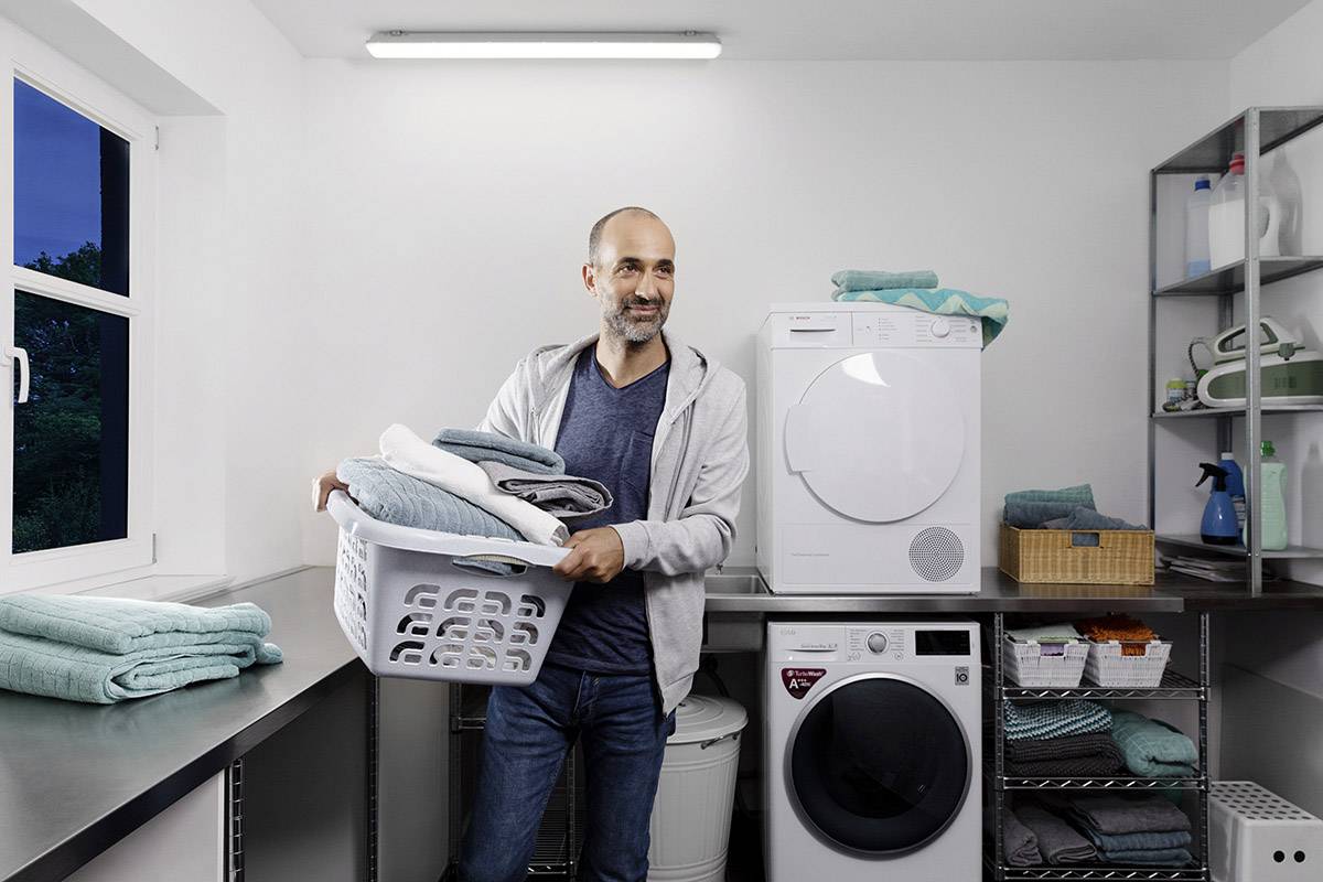 Mann in Waschküche hält Wäschekorb, steht vor Waschmaschine und Trockner. Regale mit Reinigungsmitteln und Handtüchern im Hintergrund.