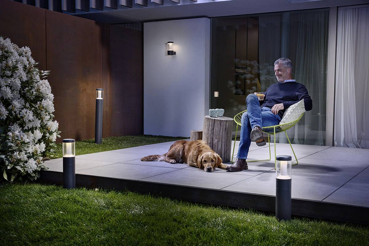 Ein Mann sitzt abends entspannt auf einer Terrasse im Garten mit einem Hund zu seinen Füßen. Moderne Beleuchtung schafft eine gemütliche Atmosphäre.