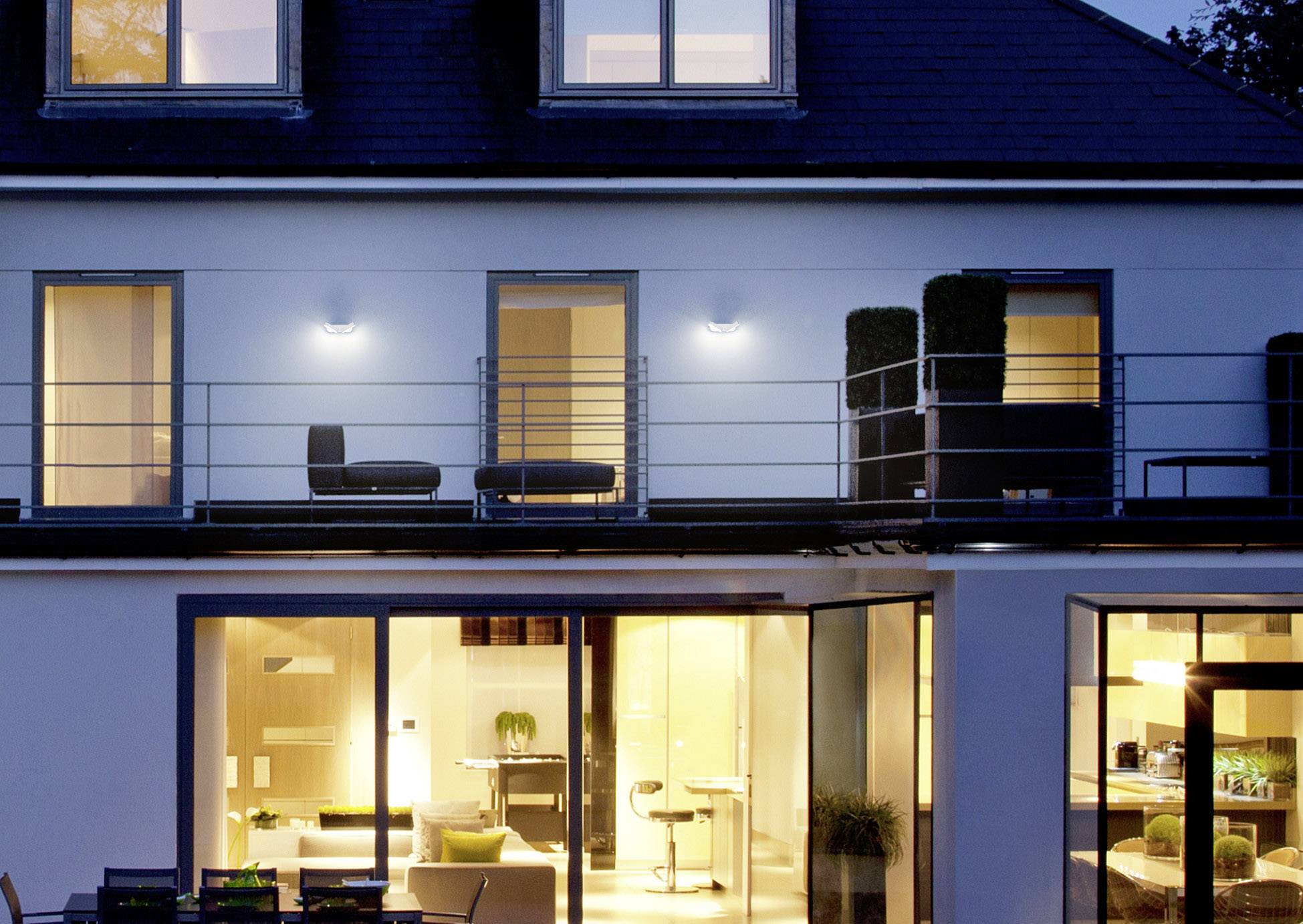 Ein modernes zweistöckiges Haus bei Nacht, beleuchtet mit großen Fenstern und Balkon im Obergeschoss. Innenbeleuchtung sichtbar.