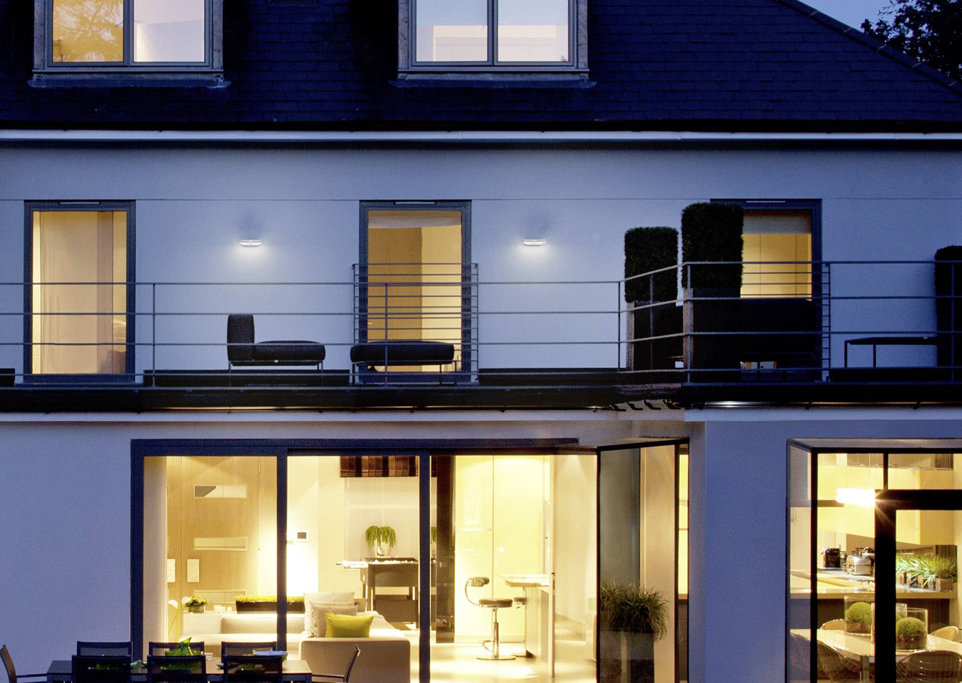 Ein modernes zweistöckiges Haus bei Dämmerung, Fenster beleuchtet. Oben Terrasse mit Möbeln, unten große Glasfront, Innenräume sichtbar.