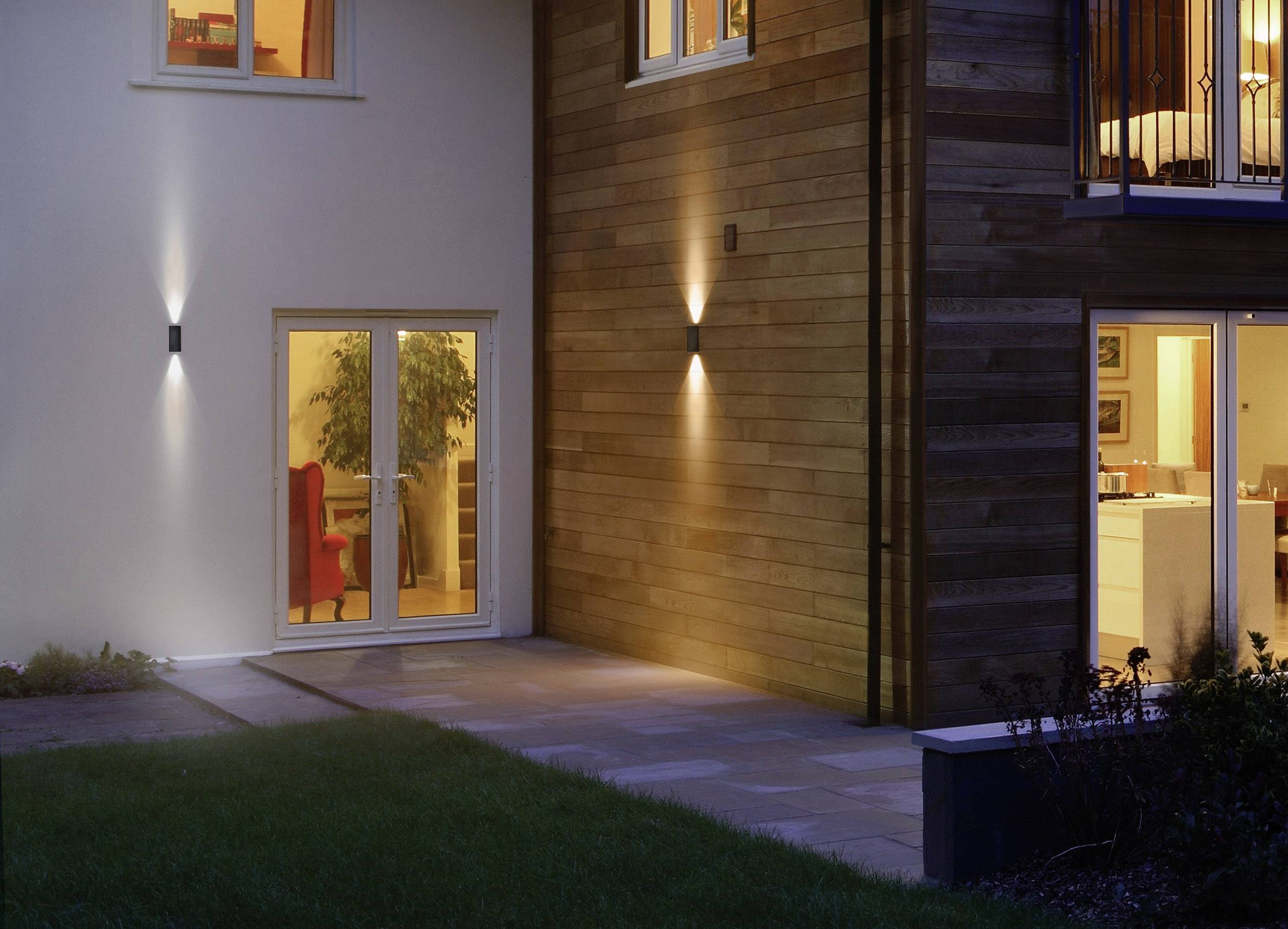 Außenansicht eines modernen Wohnhauses bei Nacht, beleuchtet durch Außenlampen. Durch die Fenster sind ein Wohnzimmer und eine Küche sichtbar.