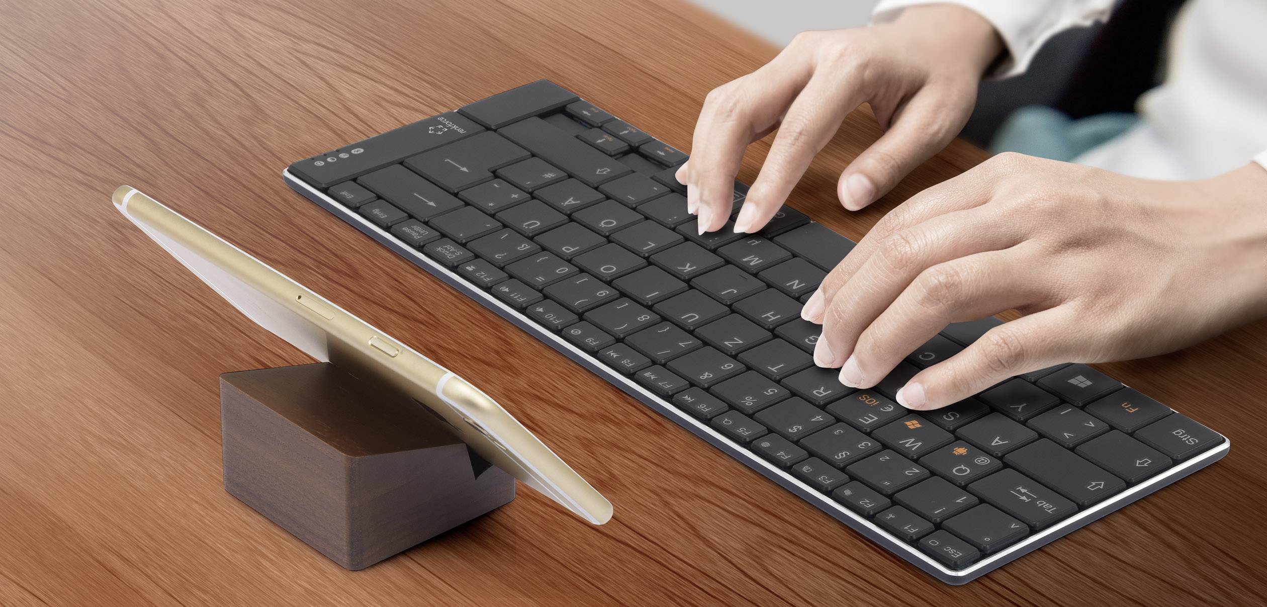 Eine Person tippt auf einer schwarzen drahtlosen Tastatur. Ein goldenes Smartphone steht neben ihr auf einem Holzständer.