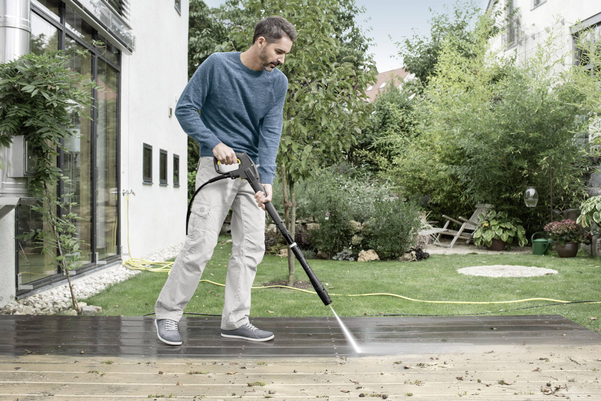Ein Mann reinigt mit einem Hochdruckreiniger eine Terrasse. Im Hintergrund sind Pflanzen und ein Haus zu sehen.