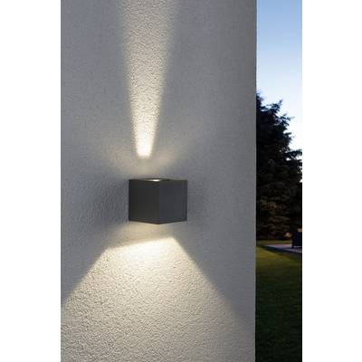Wandlampe an weißer Außenwand mit strahlendem Lichtkegel nach oben und unten. Im Hintergrund Bäume und blauer Himmel am Abend.