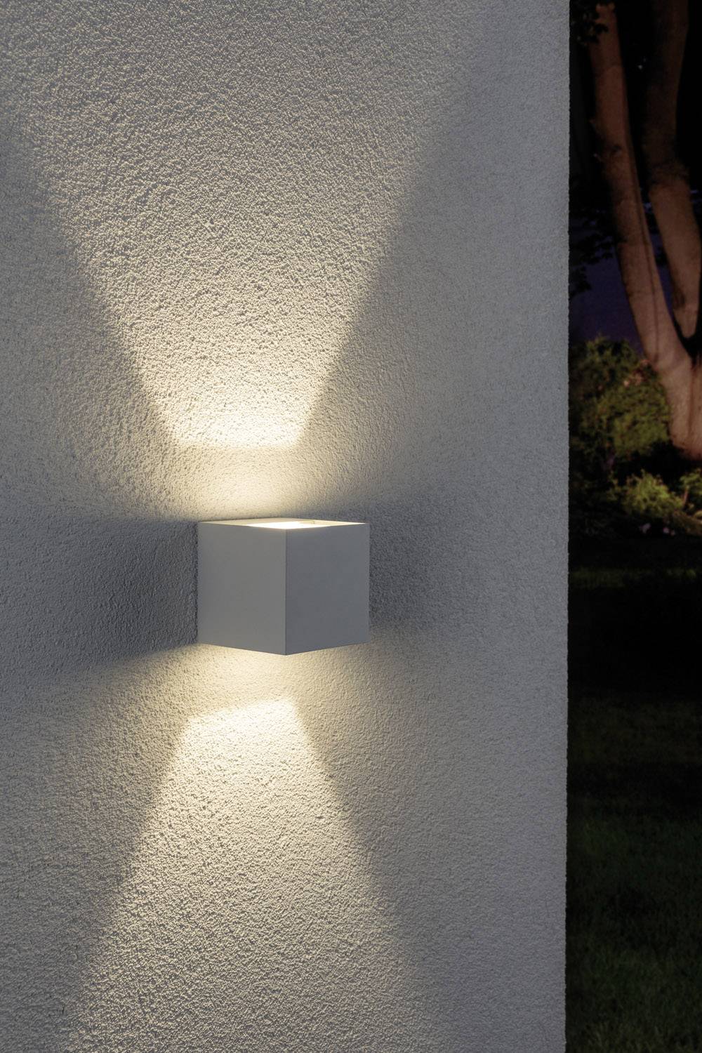 Wandleuchte wirft Lichtkegel nach oben und unten an einer strukturierten weißen Wand; im Hintergrund Bäume und Rasen.