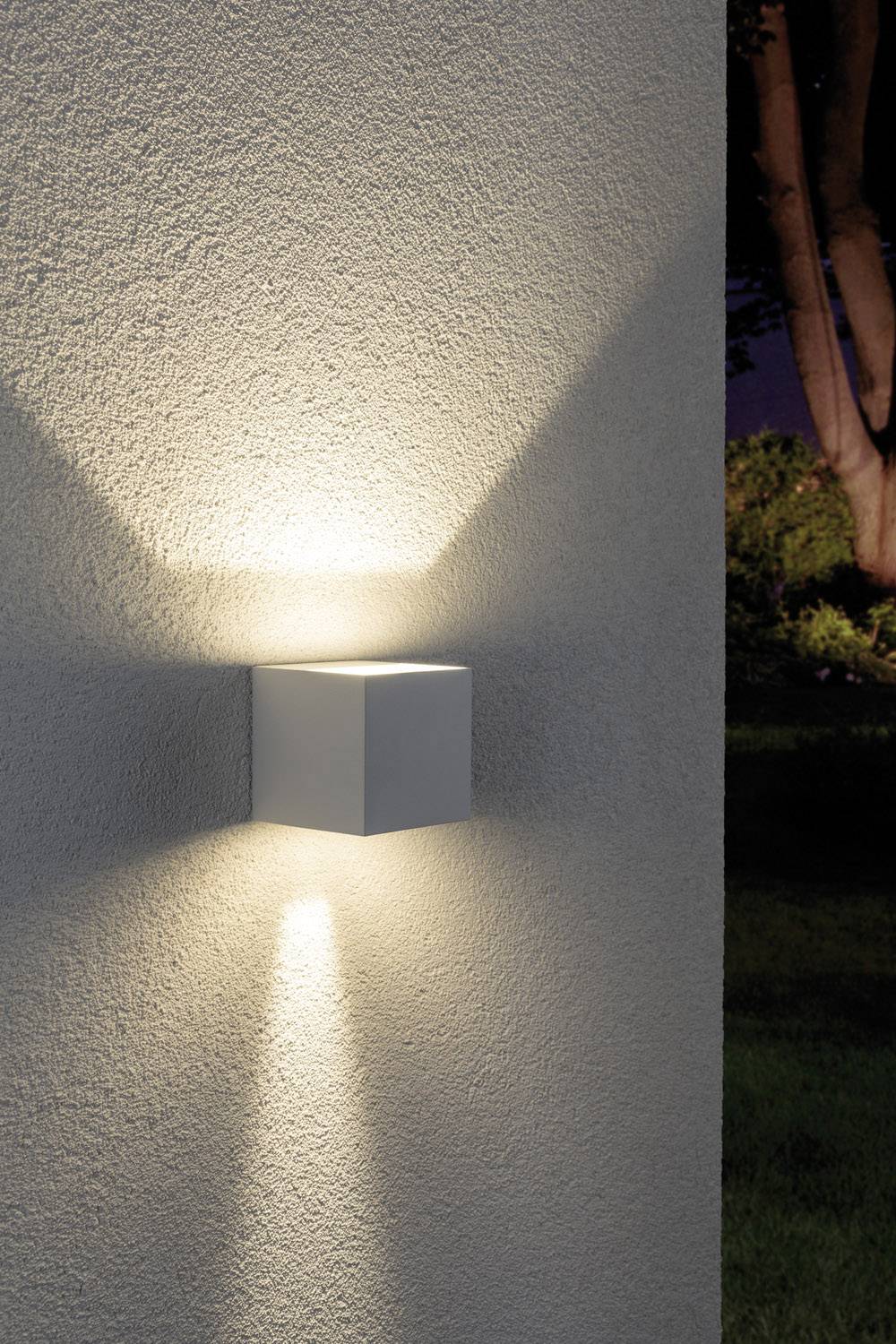 Eine moderne Wandleuchte in Würfelform an einer Hauswand. Sie strahlt Licht nach oben und unten. Hintergrund zeigt dunklen Garten.