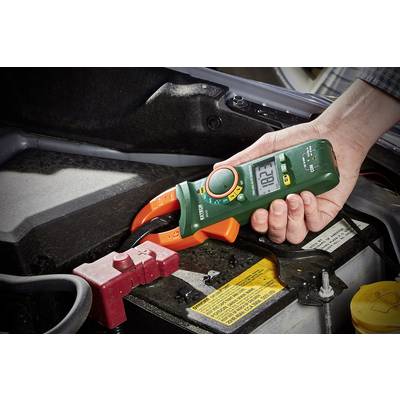 Hand hält ein digitales Multimeter und misst Spannung der Autobatterie unter der Motorhaube. Messwertanzeige zeigt '12.6'.