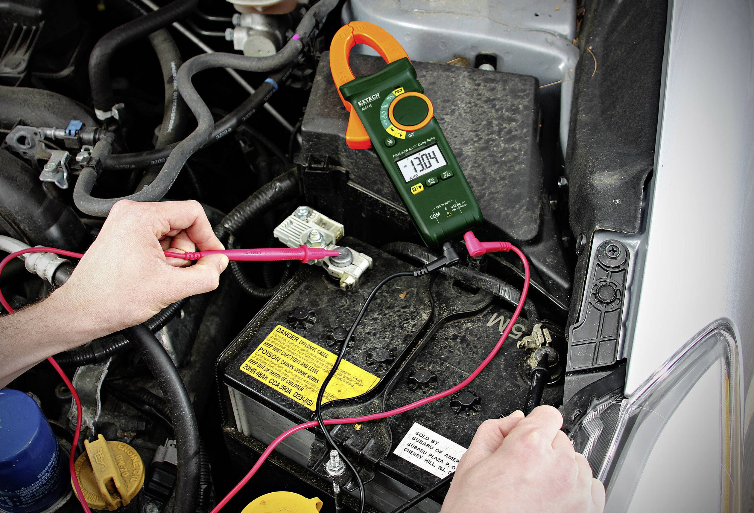 Eine Person misst die Batteriespannung eines Autos mit einem digitalen Multimeter. Der Wert auf dem Display zeigt 13,04 Volt an.