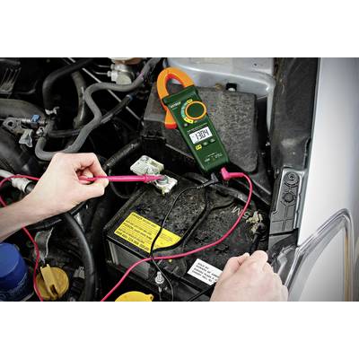 Eine Person misst die Batteriespannung eines Autos mit einem digitalen Multimeter. Der Wert auf dem Display zeigt 13,04 Volt an.