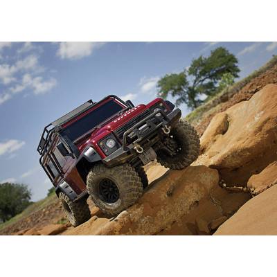 Ein rotes Geländewagen fährt über felsiges Offroad-Gelände unter blauem Himmel mit weißen Wolken. Im Hintergrund sind Bäume sichtbar.