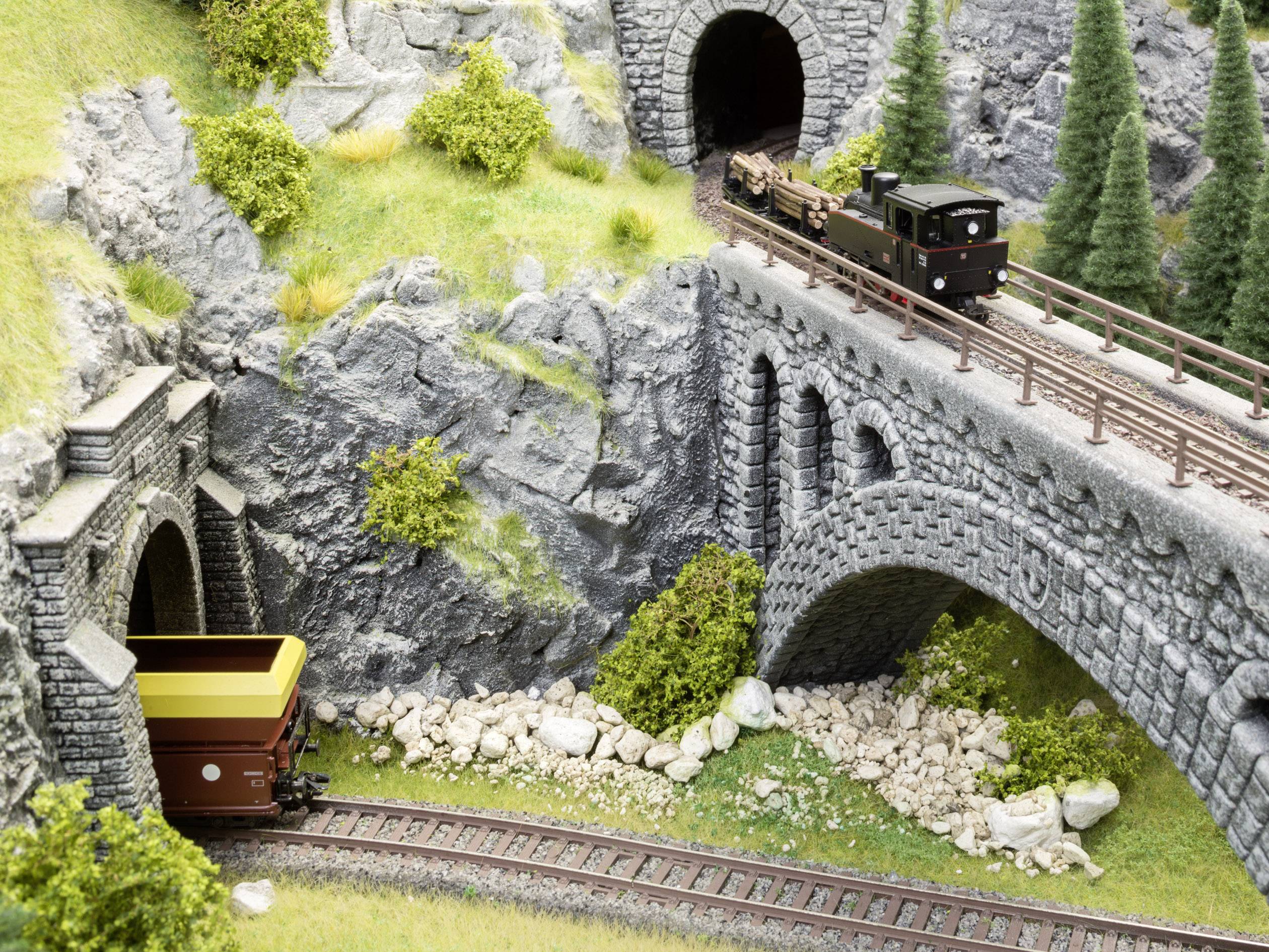 Eine Modelleisenbahn auf einem Steinviadukt, darunter ein Tunnel und ein Güterwaggon. Umliegende Landschaft mit Felsen und Bäumen.