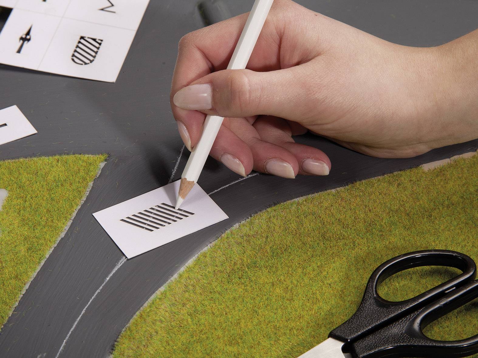 Eine Hand zeichnet mit einem Bleistift Linien auf eine kleine Karte, die auf einem Modell mit Grasflächen liegt. Daneben liegt eine Schere.