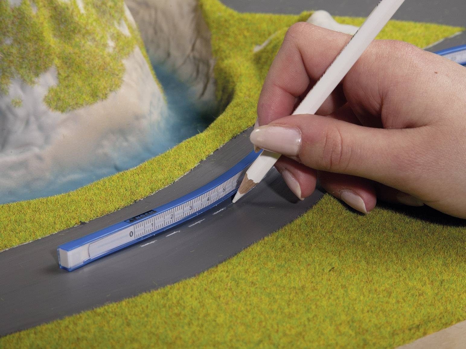 Eine Hand zeichnet mit einem weißen Bleistift Linien auf einen grauen Modellstraßenabschnitt. Verwachsene Landschaft im Hintergrund.