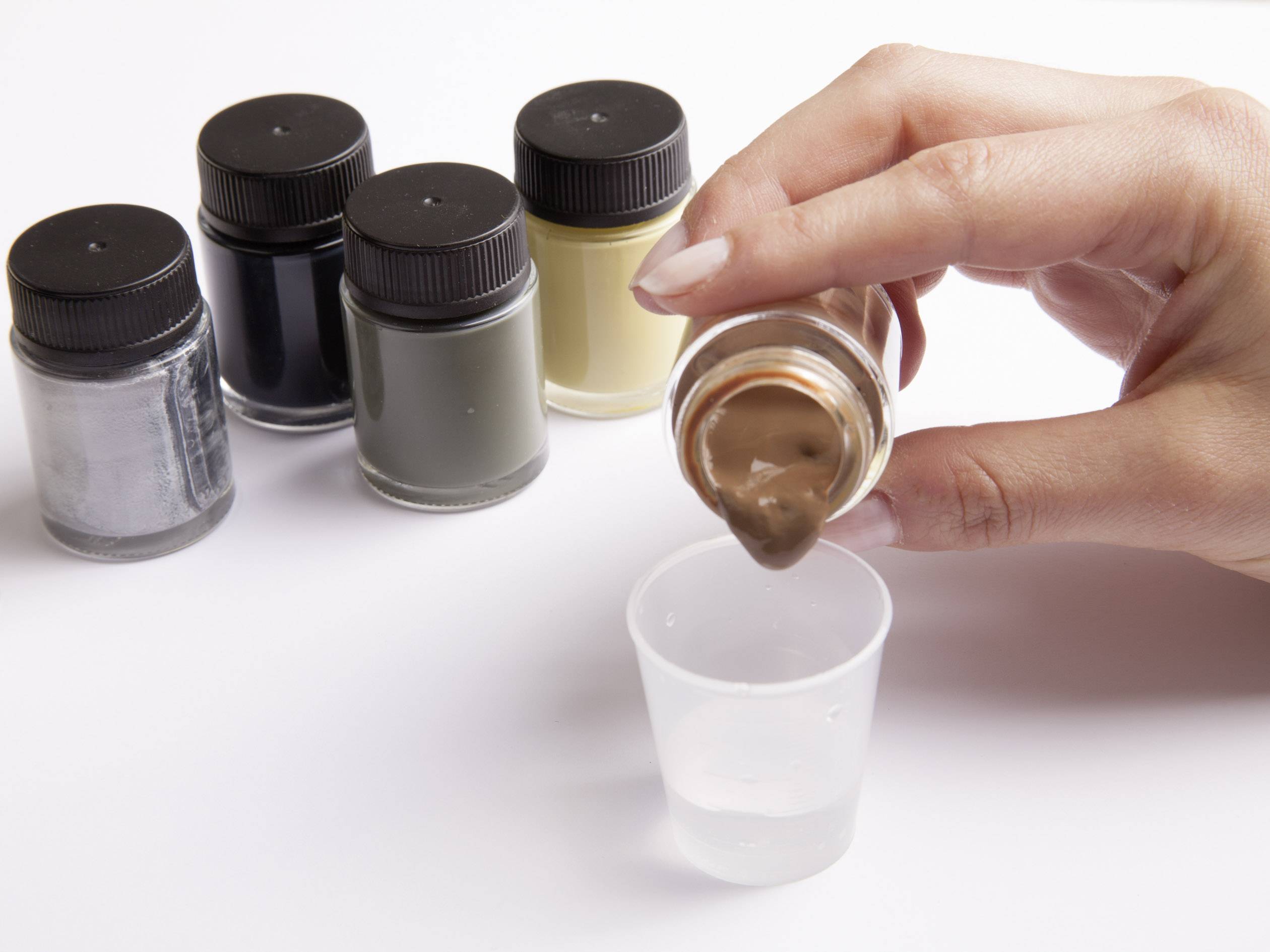 Eine Hand gießt braune Farbe aus einem Glasgefäß in einen Plastikbecher. Im Hintergrund sind vier weitere Farbgläser zu sehen.