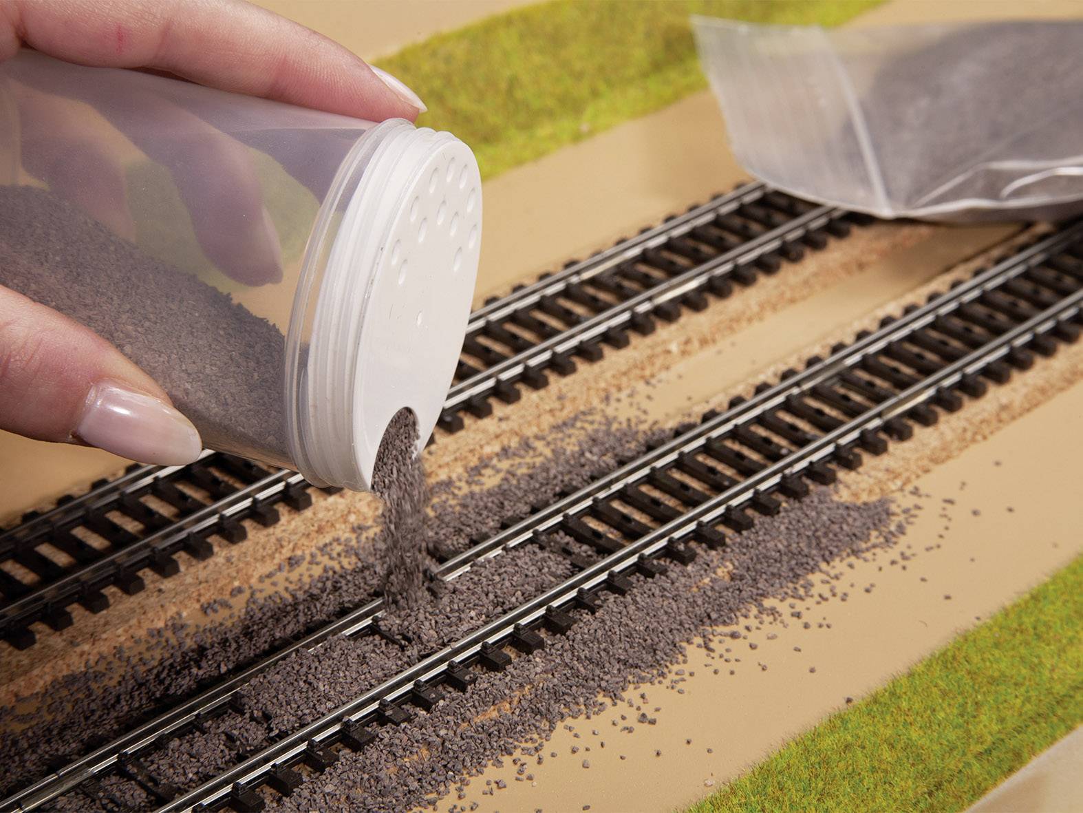 Eine Hand streut feinen grauen Kies auf ein Modellbahn-Gleisbett, um den Schotter zwischen den Gleisen zu simulieren.