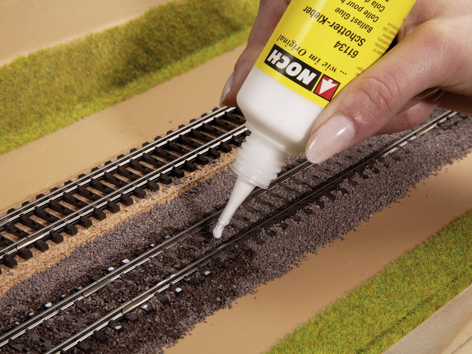 Eine Hand verwendet Kleber aus einer Flasche, um Modellbahnschienen auf einer Landschaftsplatte zu fixieren.