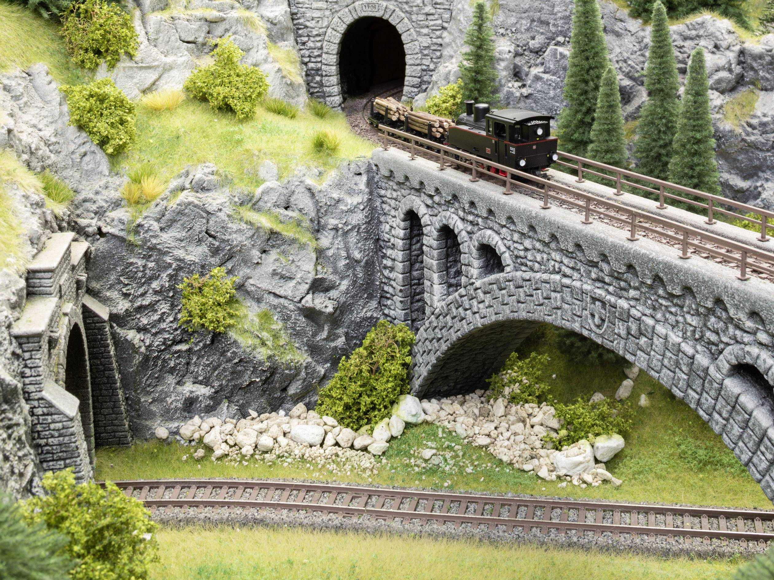 Ein Modellzug fährt über eine Steinbrücke in einer bergigen Landschaft mit grünen Bäumen und einem Tunnel im Hintergrund.