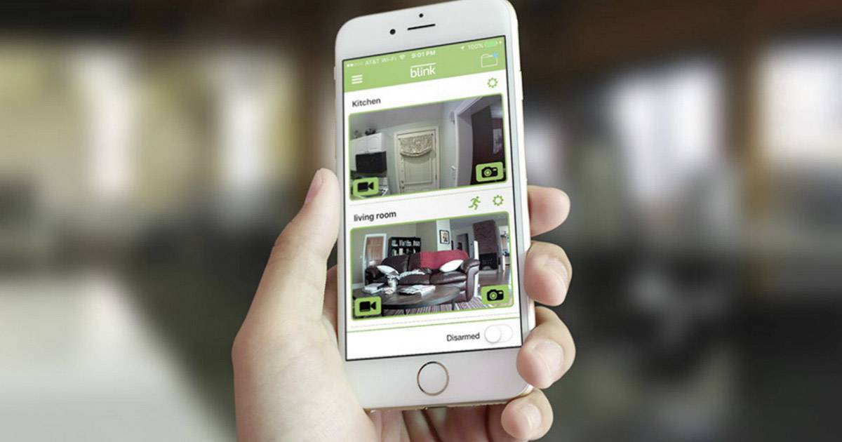 Eine Person hält ein Smartphone, das eine Überwachungs-App mit Live-Videos von 'Küche' und 'Wohnzimmer' anzeigt.