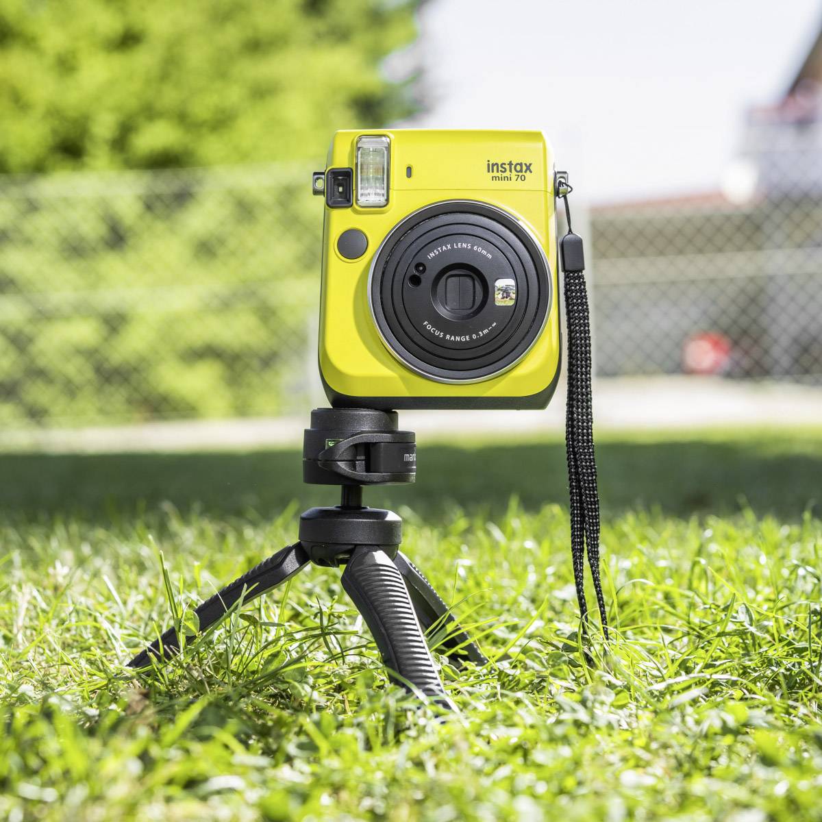 Eine gelbe Sofortbildkamera der Marke Instax Mini 70 steht auf einem Stativ im Gras, im Hintergrund unscharfe Bäume und Gebäude.