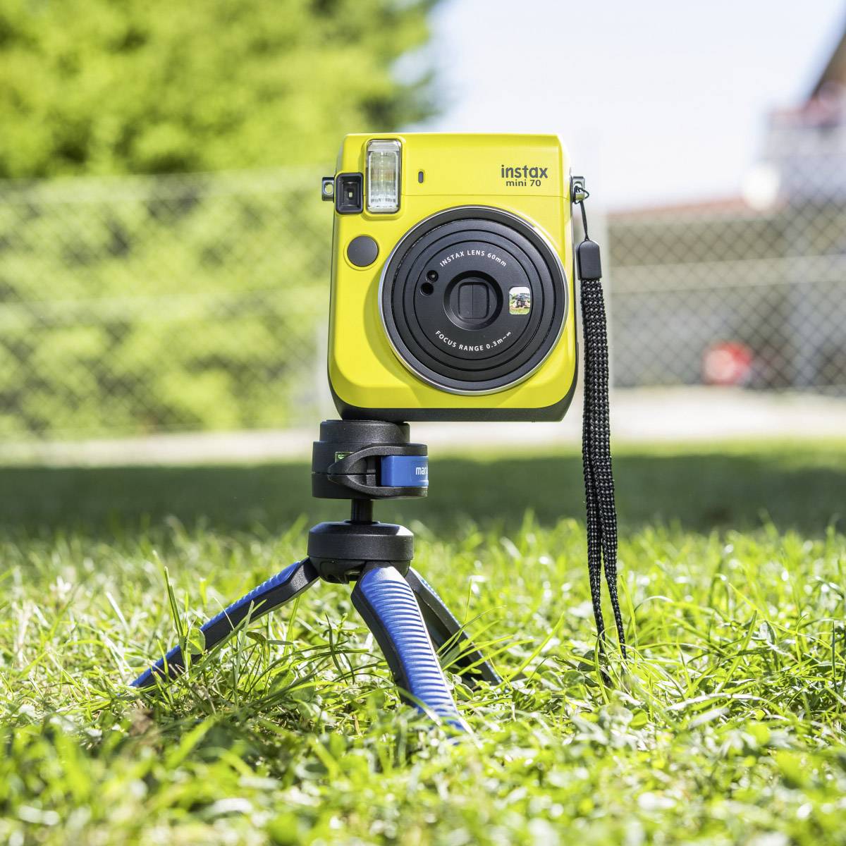 Gelbe Sofortbildkamera auf einem blauen Stativ im Freien, vor grünem Rasen und verschwommenem Hintergrund.
