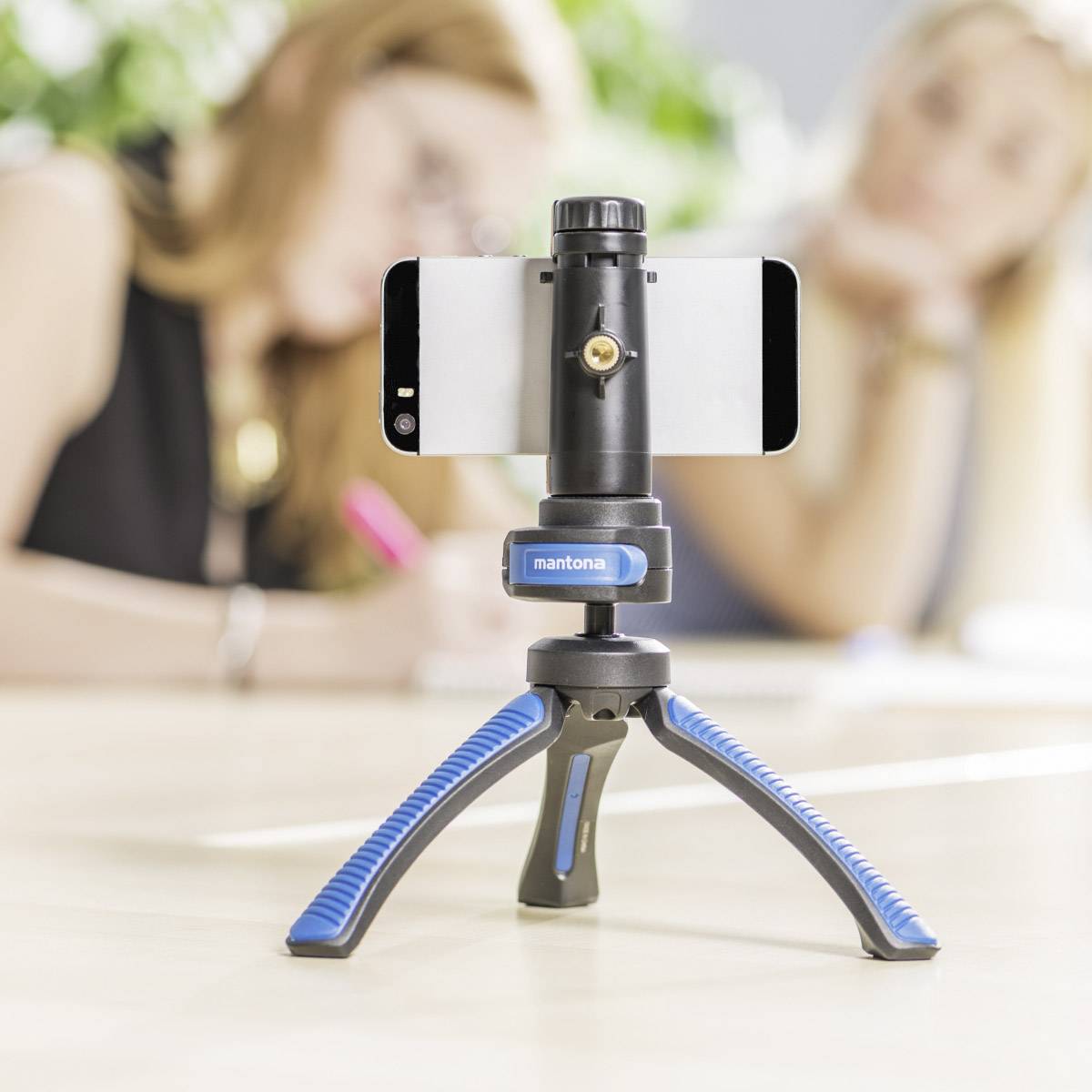 Ein Smartphone ist auf einem Stativ montiert. Zwei unscharfe Personen sind im Hintergrund, fokussiert auf das Schreiben.