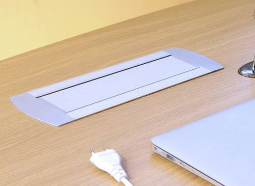 In einem Holzschreibtisch ist ein Elektrokabelschacht eingelassen, daneben liegt ein weißes Netzkabel und ein Laptop.