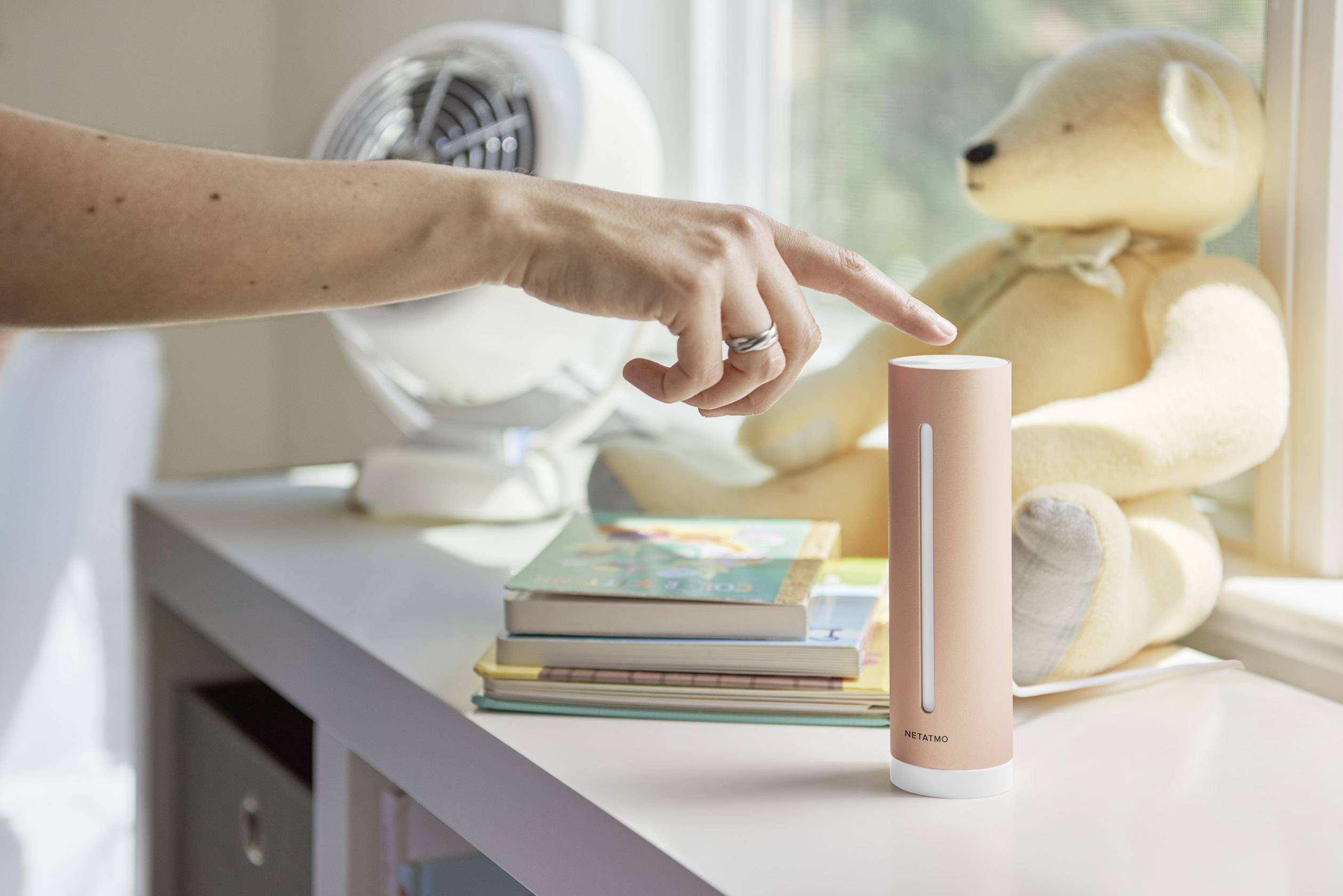 Eine Hand bedient einen zylindrischen Smart-Home-Sensor auf einem Tisch. Im Hintergrund sind ein Ventilator, Bücher und ein Stoffbär.
