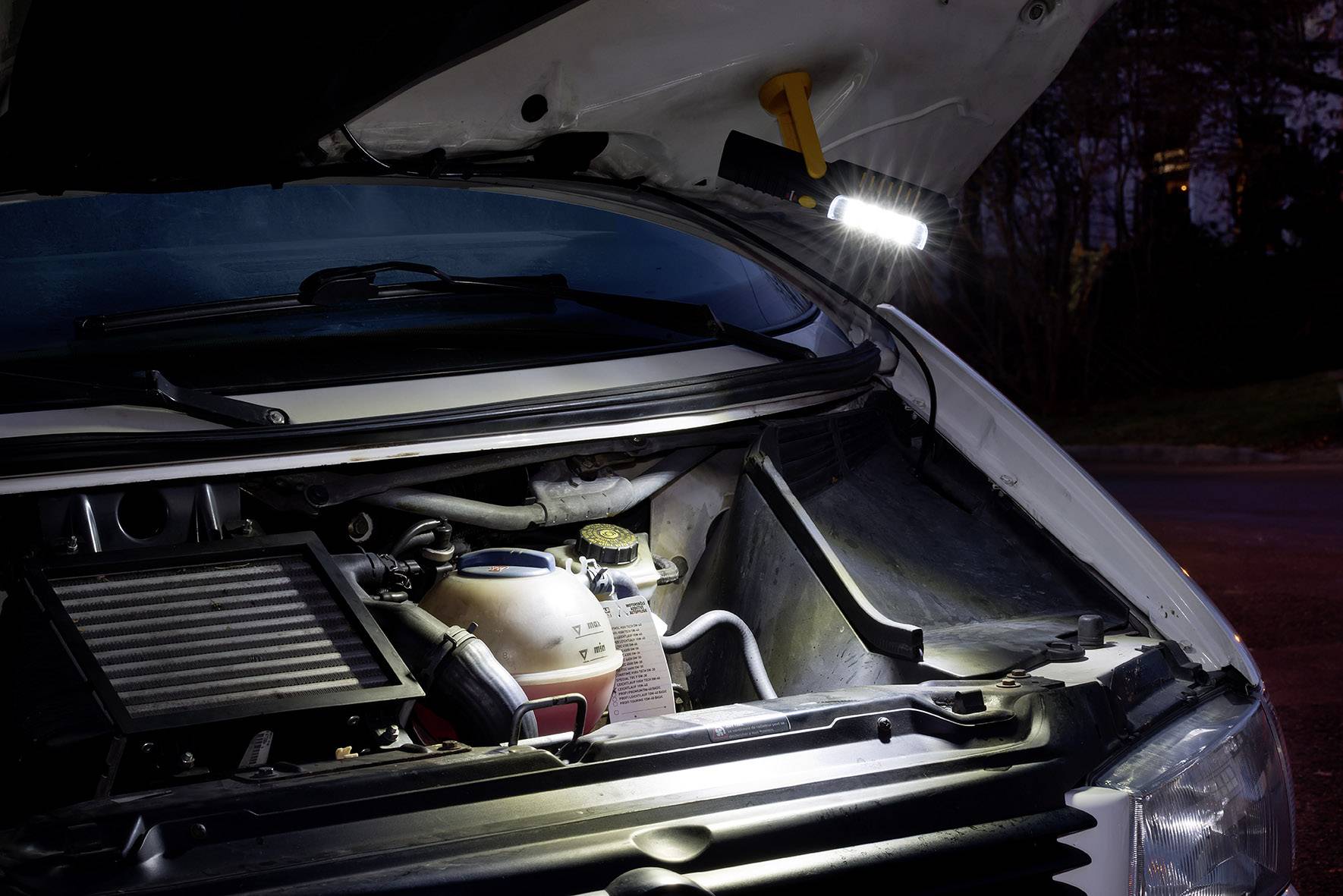 Motorhaube eines Fahrzeugs bei Nacht geöffnet; eine Taschenlampe beleuchtet den Motorraum, der ein Kühlmittelbehälter und andere Teile zeigt.