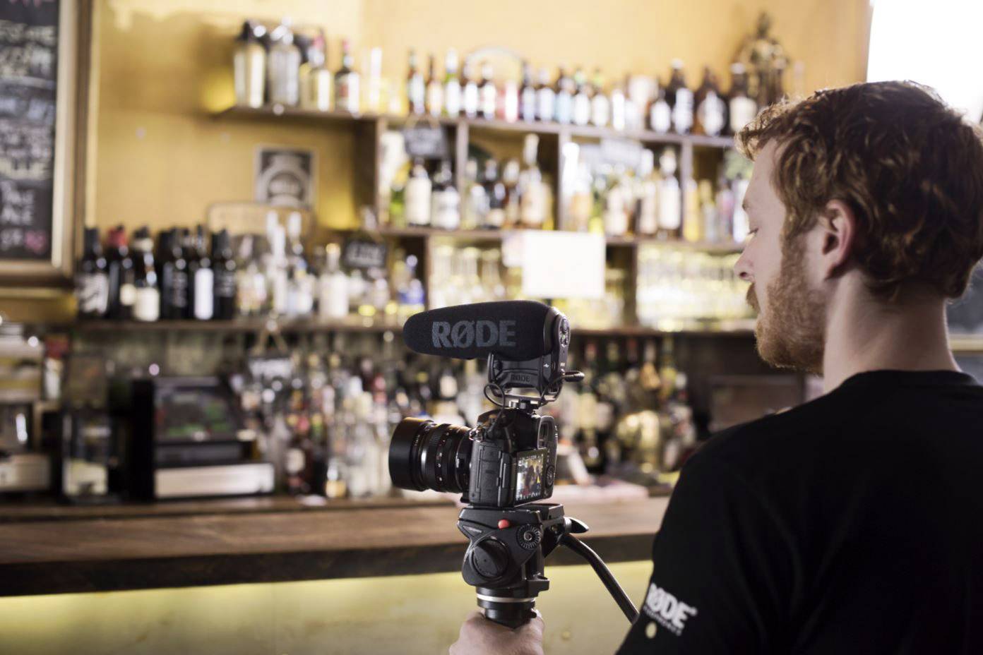 Ein Mann filmt mit einer Kamera in einer stilvollen Bar, Regale voller Flaschen im Hintergrund. Er konzentriert sich auf seine Aufnahme.