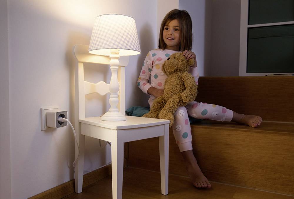 Ein Kind sitzt in Schlafanzug auf einer Treppe und hält einen Plüschbären. Neben ihr steht ein kleiner Tisch mit Lampe.