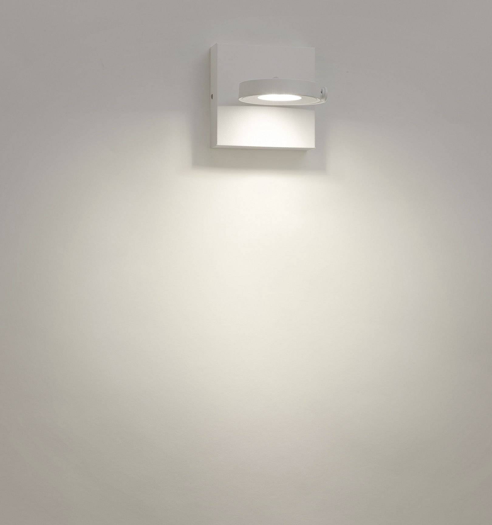 Eine schlichte Wandlampe mit rechteckigem Gehäuse aus weißem Metall leuchtet mit einem weichen, breiten Lichtstrahl nach unten.