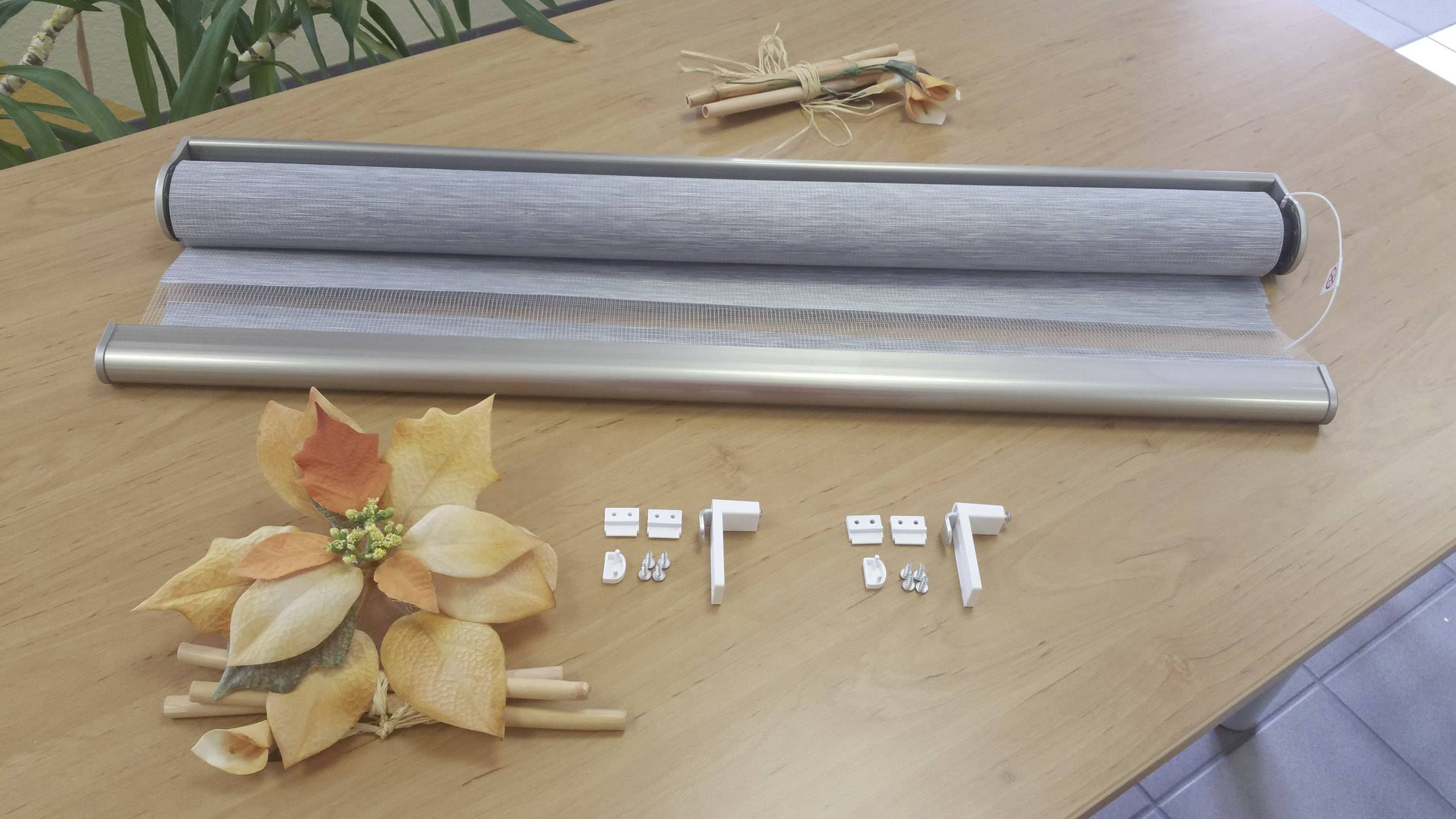 Zwei Rollos auf einem Tisch mit Montagezubehör. Links liegt herbstliche Dekoration aus Kunstblättern. Im Hintergrund Pflanzen.