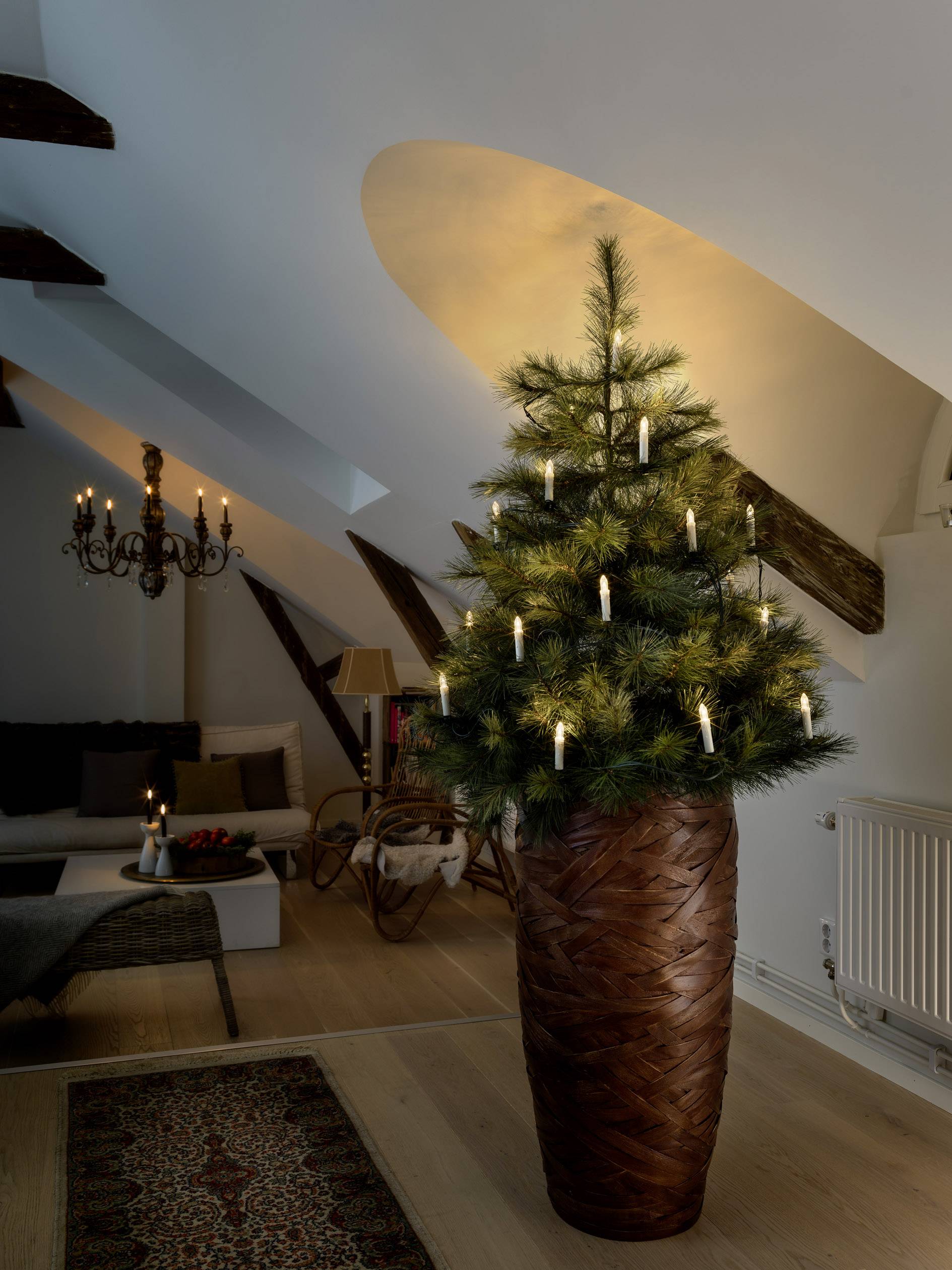 Ein geschmückter Tannenbaum in einem gemütlich beleuchteten Wohnzimmer mit Kerzen und einem Kronleuchter an der Decke.