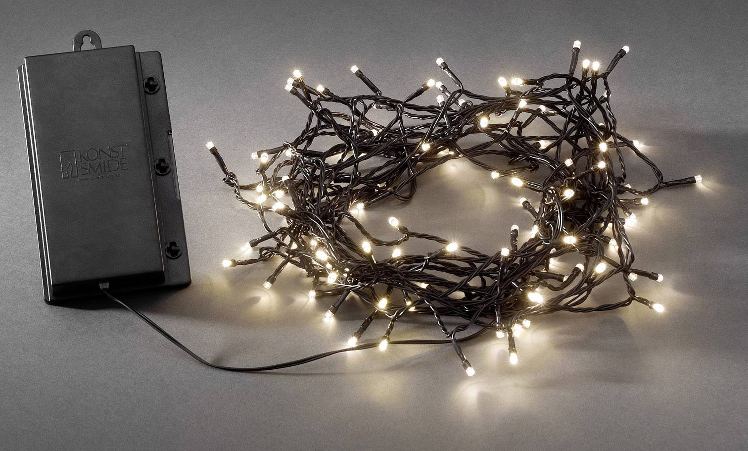 Eine Lichterkette mit kleinen, leuchtenden Glühbirnen liegt neben einem schwarzen Batteriefach auf grauem Hintergrund.