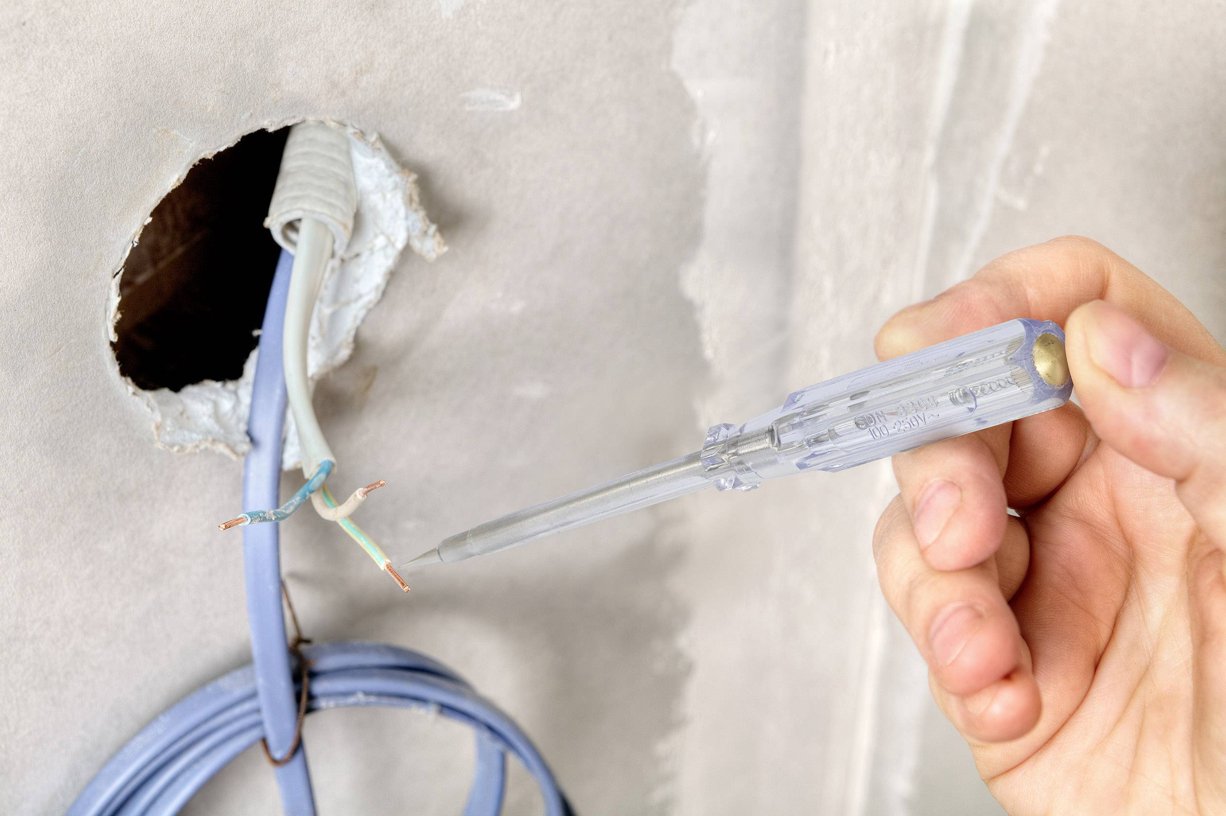 Person hält einen Spannungsprüfer und überprüft freiliegende Drähte, die aus einem Loch in der Wand herausragen.