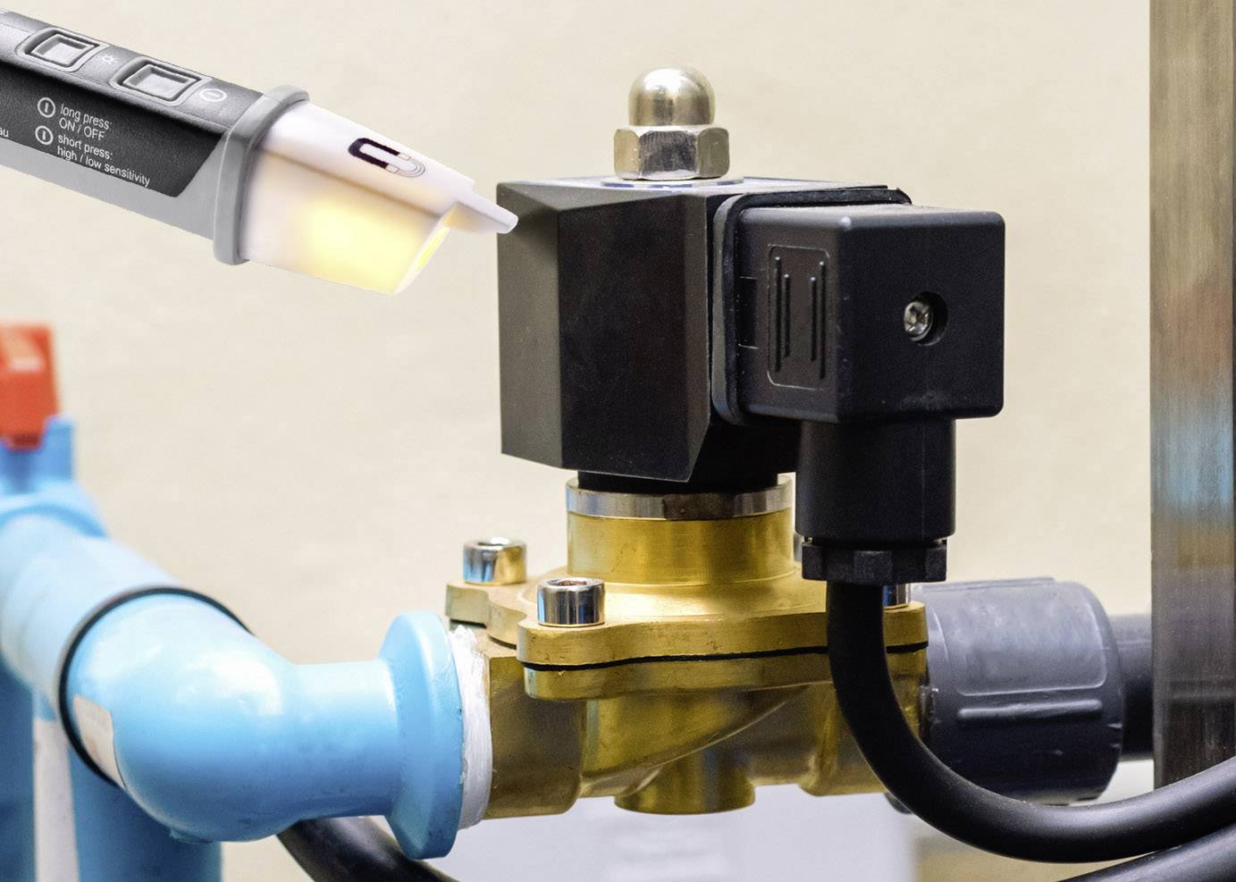 Ein blaues Rohr mit einem goldfarbenen Ventil und einem schwarzen elektrischen Bauteil verbunden. Eine Hand hält ein Prüfgerät an das Ventil.