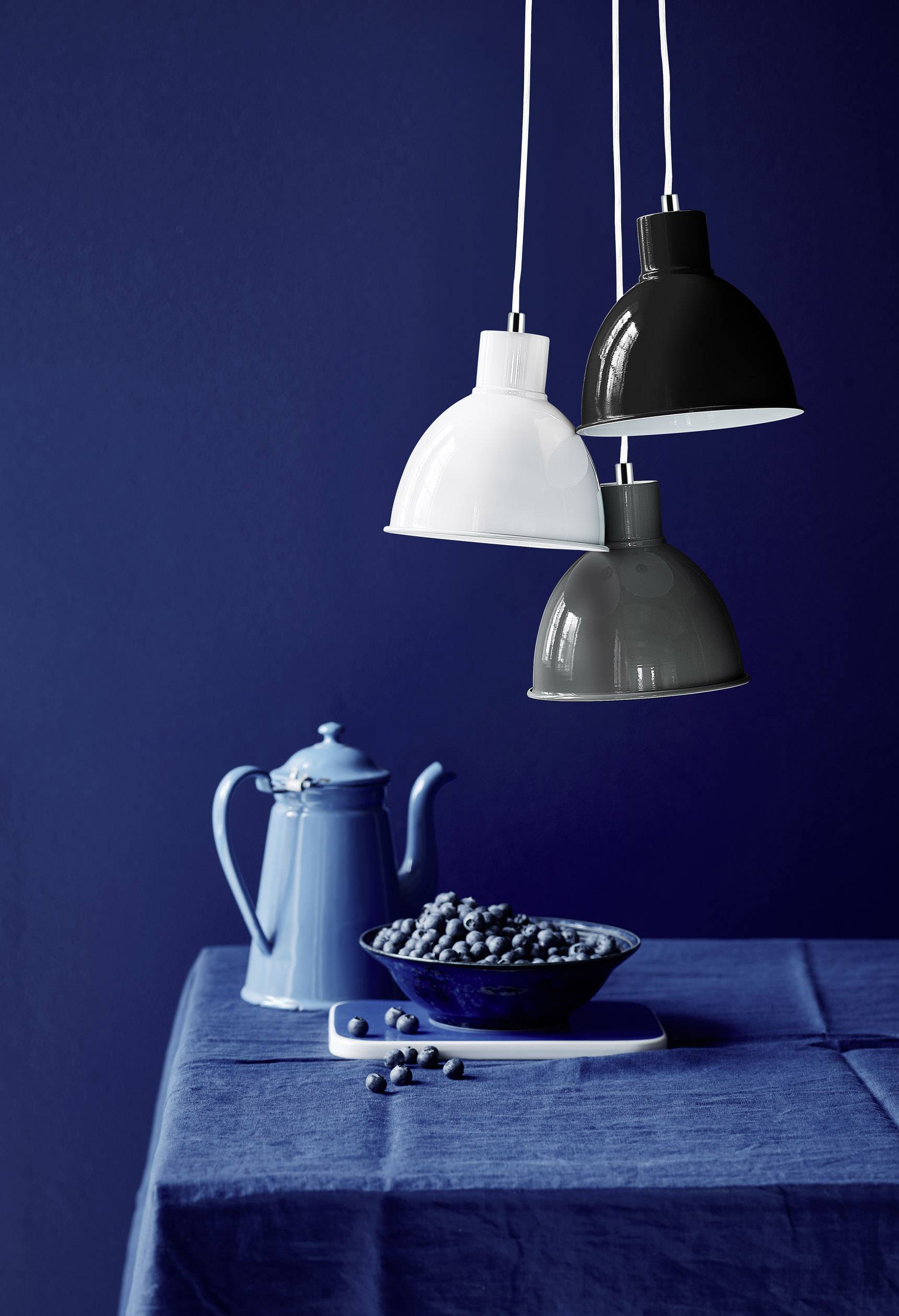 Drei hängende Lampen in Weiß, Schwarz und Grau über einem Tisch mit einer blauen Kanne und einer Schale voller blauer Früchte.