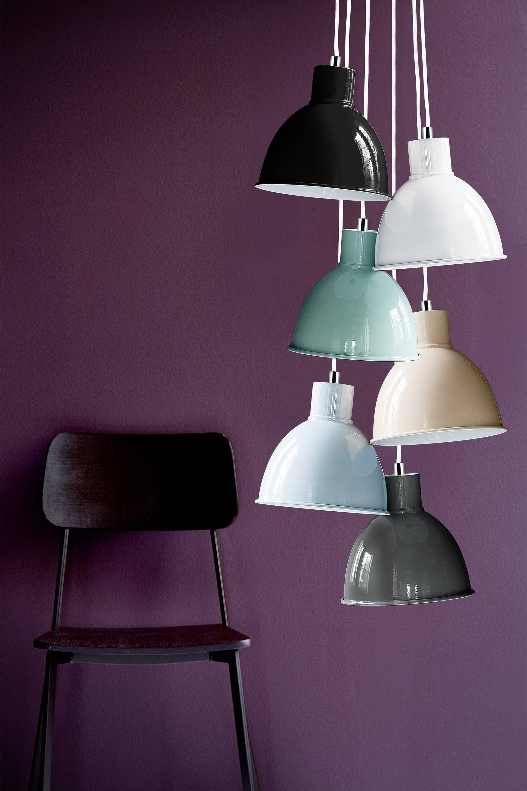 Sechs unterschiedlich farbige Hängelampen, darunter schwarz, weiß, grün, beige, blau und grau, hängen vor einer dunkelvioletten Wand neben einem Stuhl.