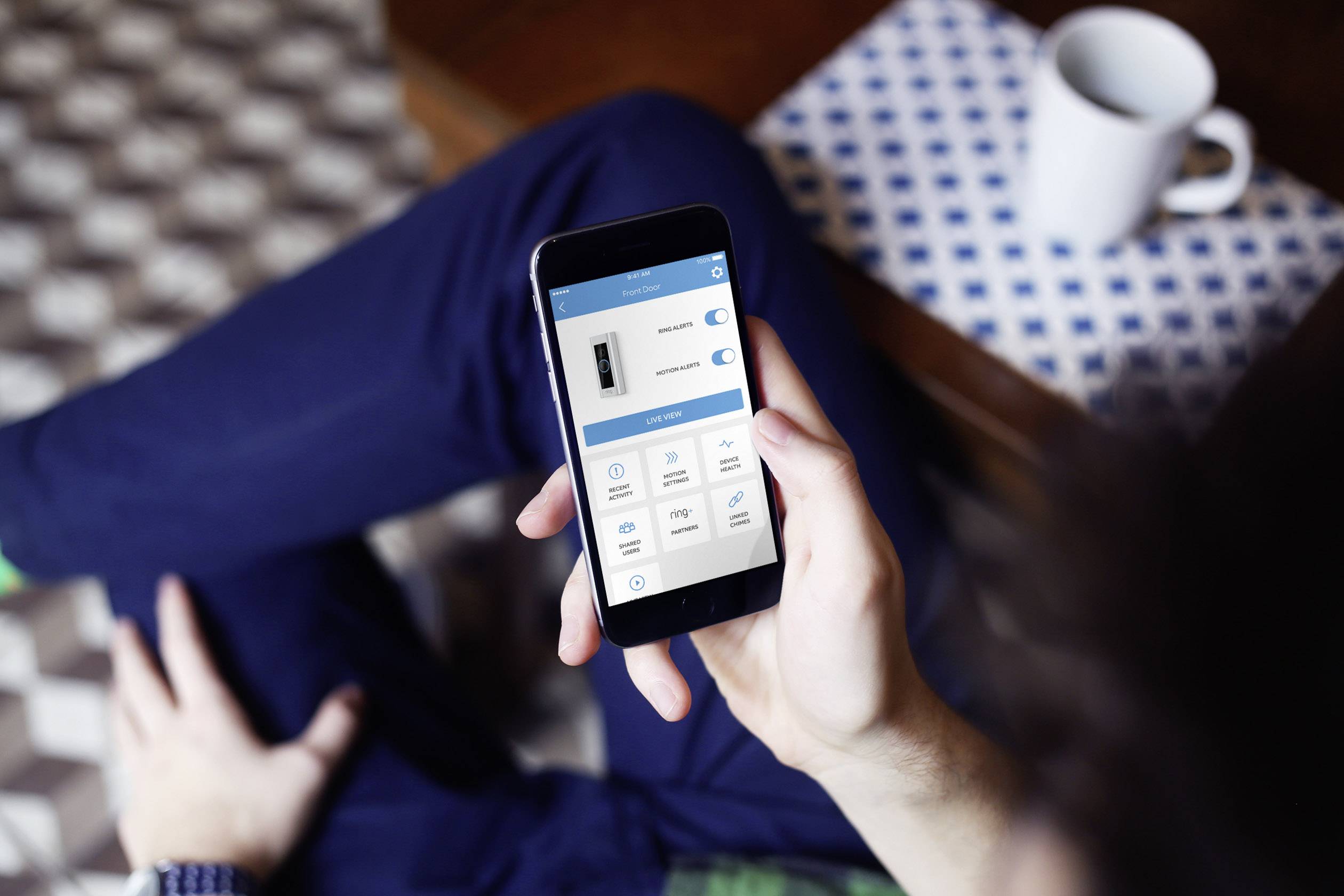 Person hält ein Smartphone mit einer geöffneten App. Die App zeigt Schalter und Optionen zur Steuerung eines Geräts. Tasse im Hintergrund.