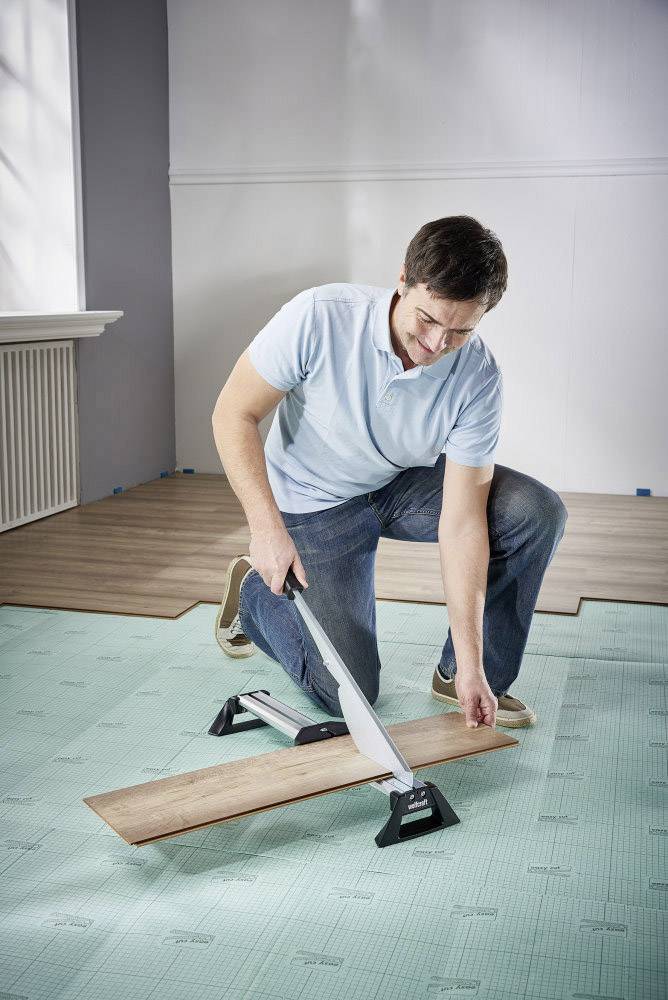 Ein Mann schneidet Laminatboden mit einer Säge in einem Raum. Hinter ihm sind bereits teilweise verlegte Laminatdielen zu sehen.