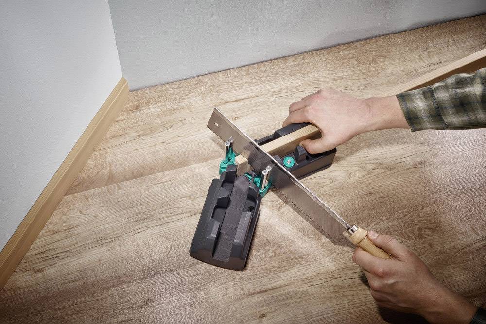 Eine Person verwendet eine Säge und eine Gehrungslade, um eine Holzleiste im 45-Grad-Winkel zu schneiden. Der Boden besteht aus hellem Holz.