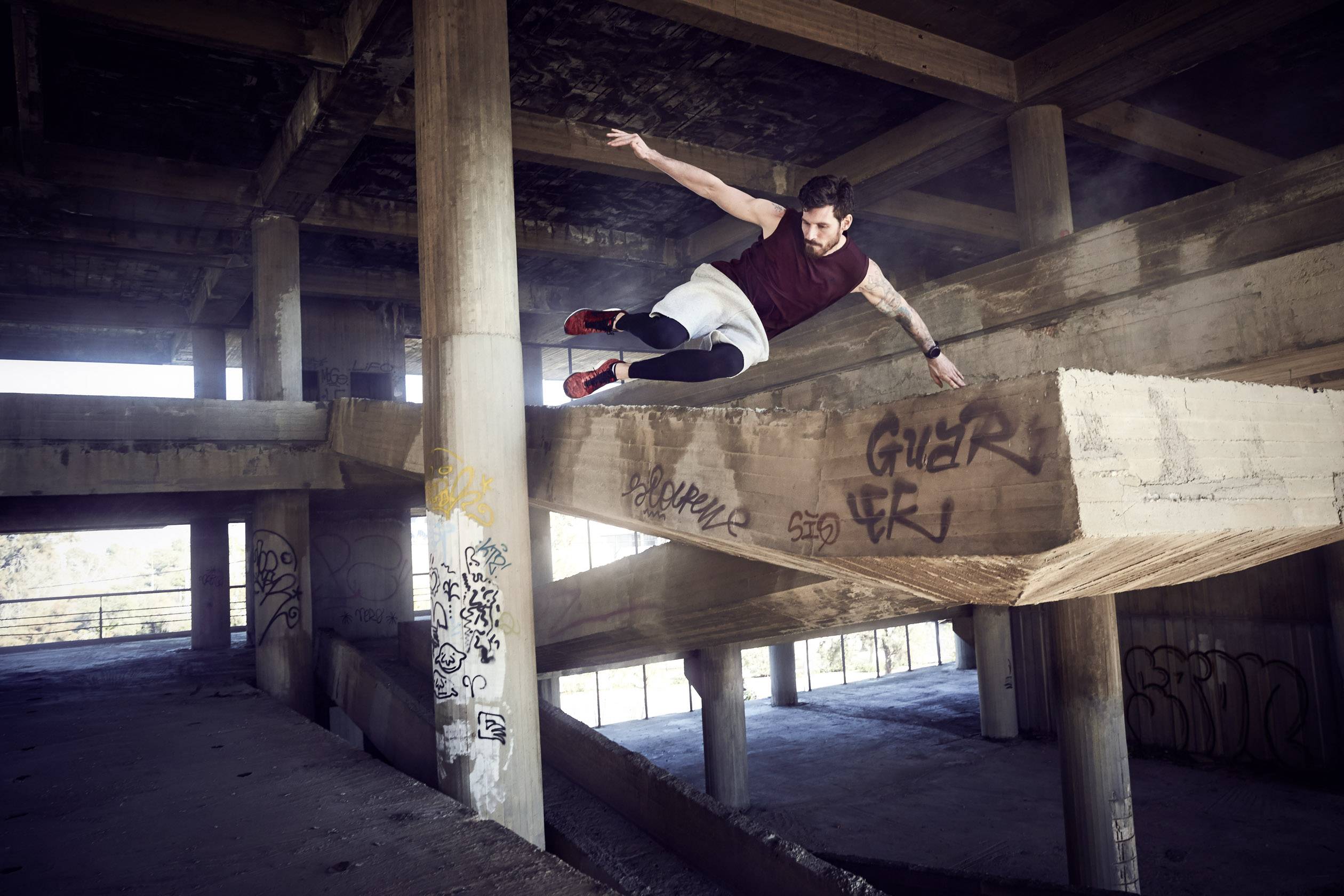 Ein Mann führt Parkour in einem verlassenen Betongebäude durch, während er über eine Wand springt. Grafitti sind auf der Wand sichtbar.