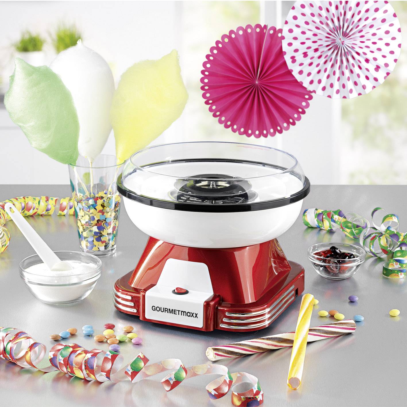 Eine Zuckerwattemaschine auf einem Tisch, umgeben von buntem Zucker, Zuckerwattestäben, Sprinkles und Dekorationen.