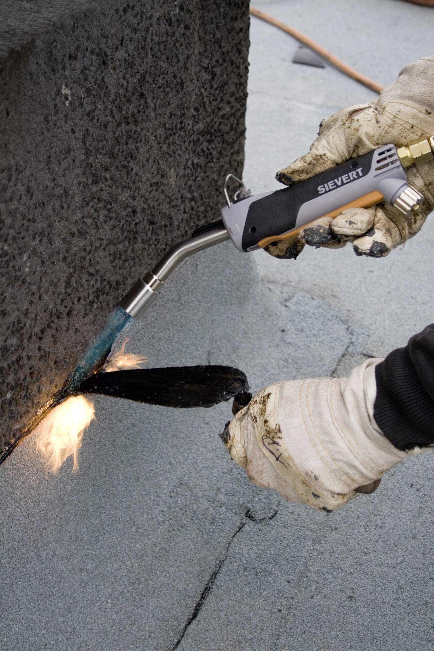 Person mit Handschuhen bedient eine Flamme mit einem Werkzeug, um Dachmaterial zu erhitzen und zu versiegeln.