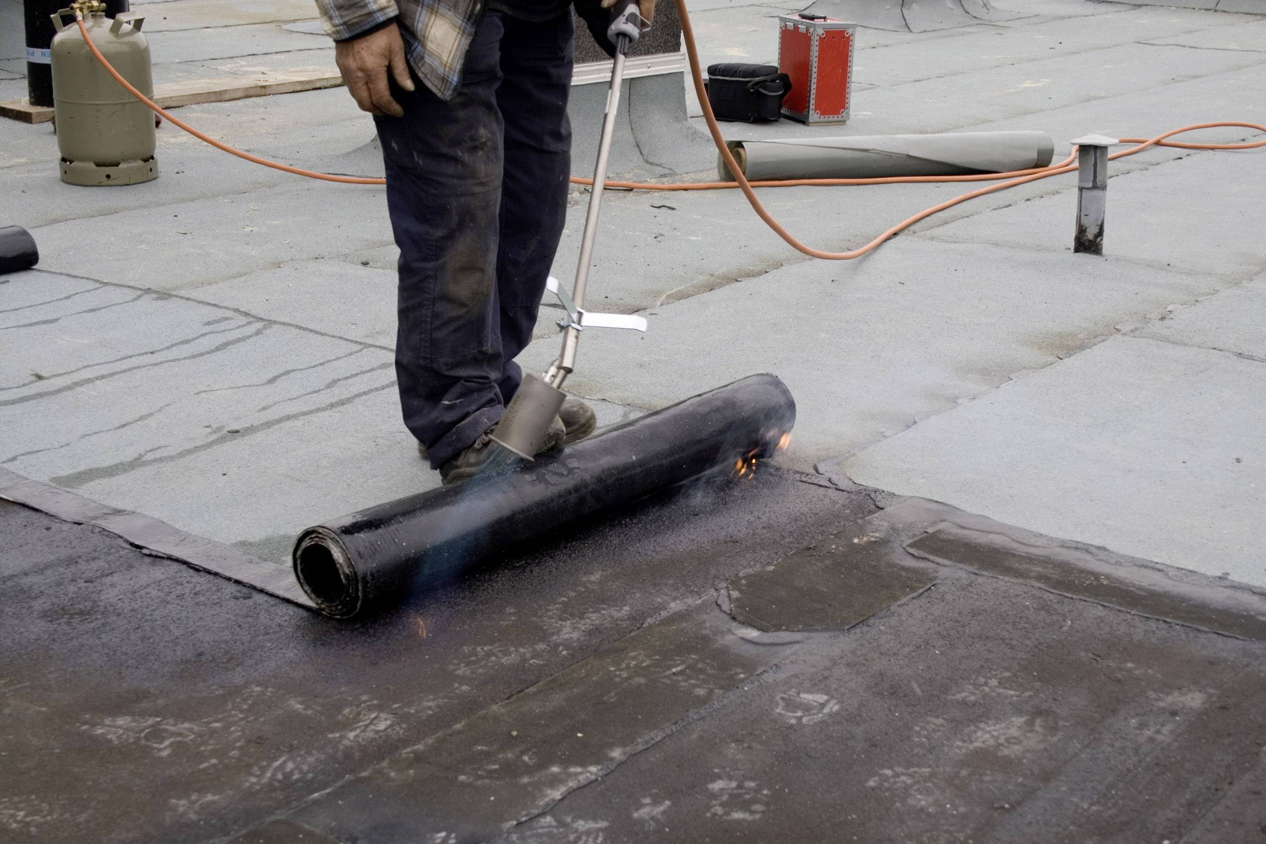 Eine Person arbeitet auf einem Dach, verwendet einen Gasbrenner, um Dachbahnen zu verschweißen. Es sind Arbeitsmaterialien sichtbar.