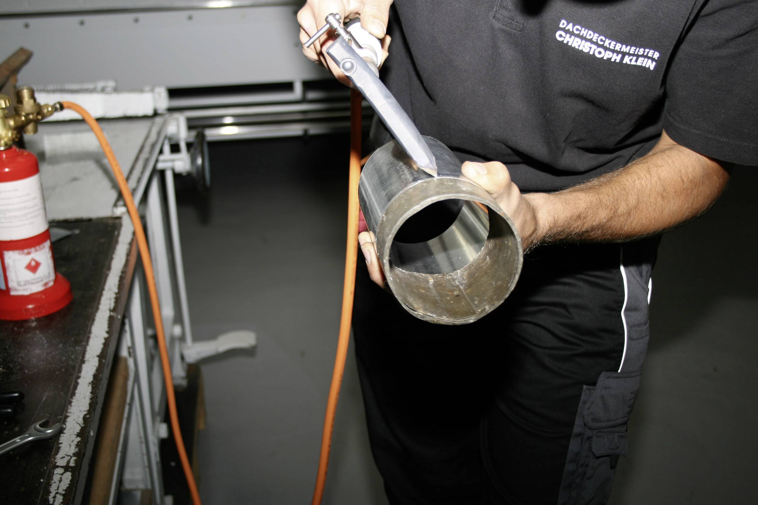Eine Person hält und bearbeitet ein Metallrohr in einer Werkstatt. Auf der Arbeitskleidung steht 'Dachdeckermeister'.